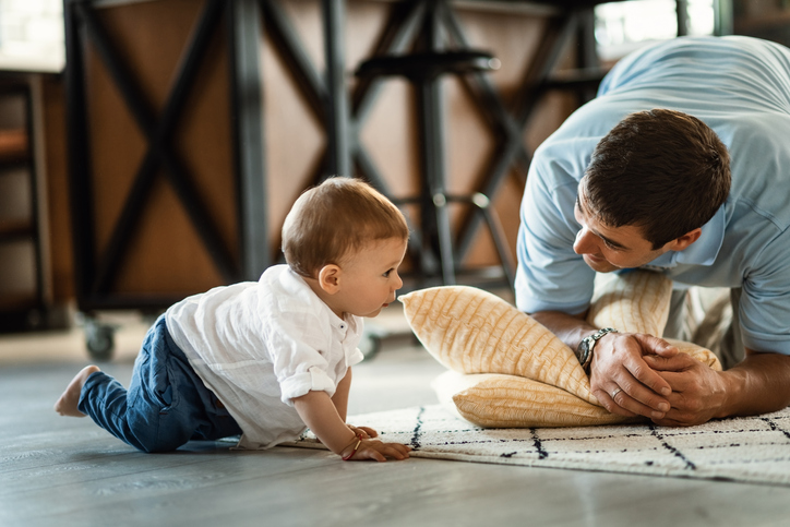 Happy father talking to his small son who is crawling on the floor in the living room.