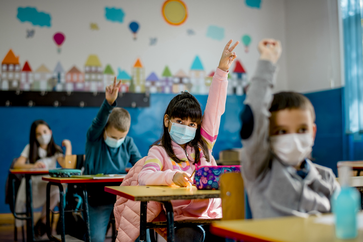 Kids in class with masks, raising their hands.