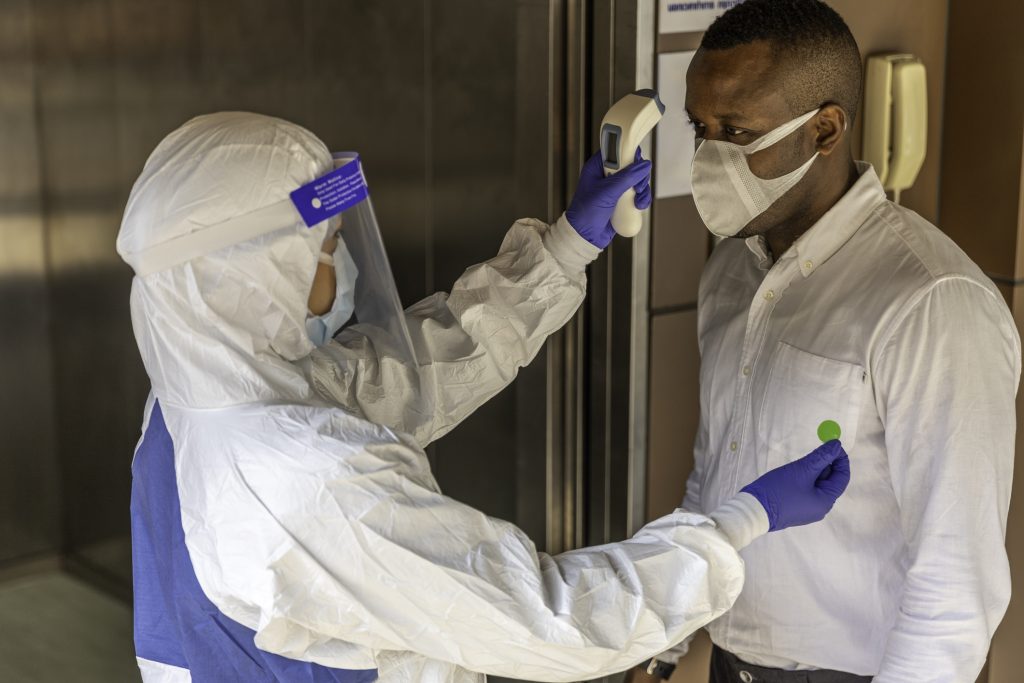 Nurse in PPE suit uses infrared thermometer measuring temperature with African male worker scanning for Coronavirus or Covid-19 symptom at elevator 