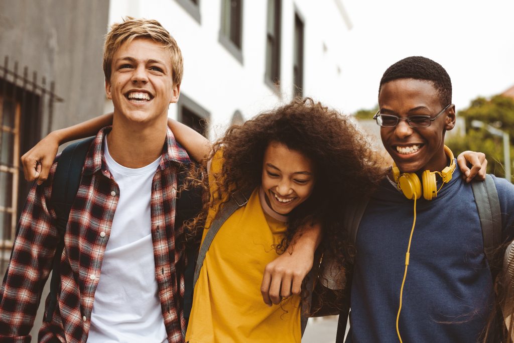 Close up of three college friends standing in the street with arms around each other. Cheerful boys and a girl wearing college bags having fun walking outdoors.