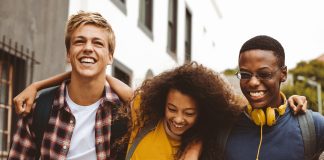 Close up of three college friends standing in the street with arms around each other. Cheerful boys and a girl wearing college bags having fun walking outdoors.